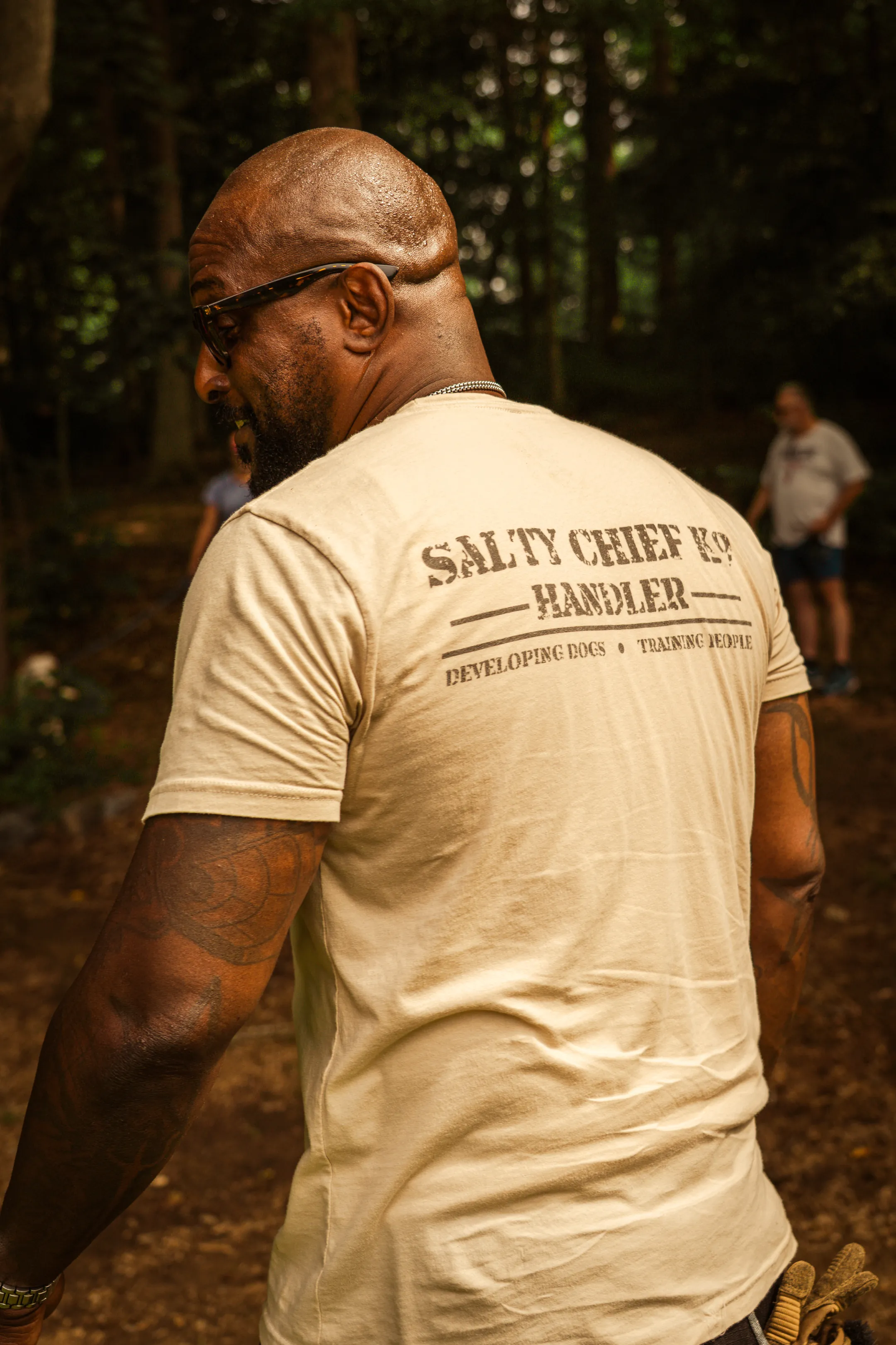 Jay in the Salty Chief K-9 handler shirt, its Developing Dogs, Training People mark across the back, turning to look over his shoulder on a wooded trail.